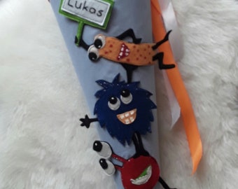 May include: A blue and grey school cone decorated with cartoon monsters and the name "Lukas". The cone has a blue star pattern at the top and bottom, with a red trim and orange ribbon.