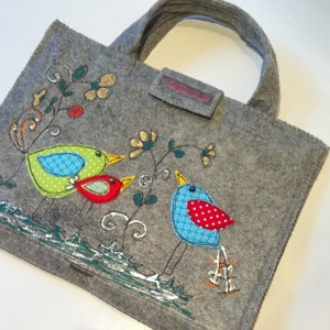 Könnte beinhalten: Graue Filz-Tragetasche mit einem skurrilen Vogeldesign. Die Vögel sind in Grün-, Rot- und Blautönen gehalten, mit dekorativen floralen Akzenten. Die Tasche hat einen Griff und ein kleines Etikett.