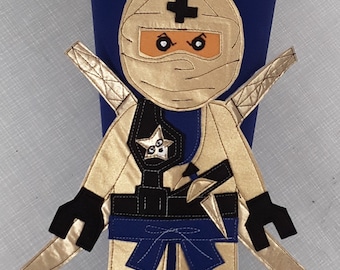 Może przedstawiać: Niebieska torba w kształcie stożka z aplikacją ninja. Ninja jest złoty, czarny i niebieski, z gwiazdą na piersi. Imię "Matty" jest wyszyte złotem. Górna i dolna krawędź mają złote wykończenia.