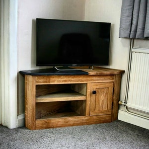 May include: A corner TV stand made of wood with a black top and a door. The stand has two shelves and a black door knob. The stand is in a room with a white wall and a grey curtain.