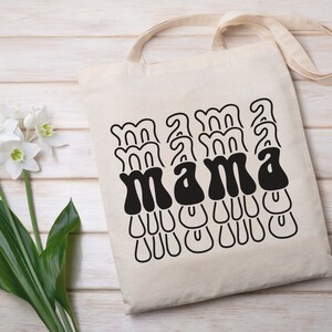 May include: A white canvas tote bag with black text that reads "mama" in a retro, wavy font.