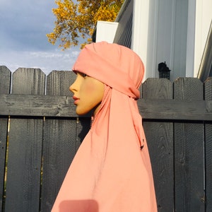 May include: A pink hijab with a decorative stitch around the edges. The hijab is draped over a mannequin head and is styled with a loose, flowing look.