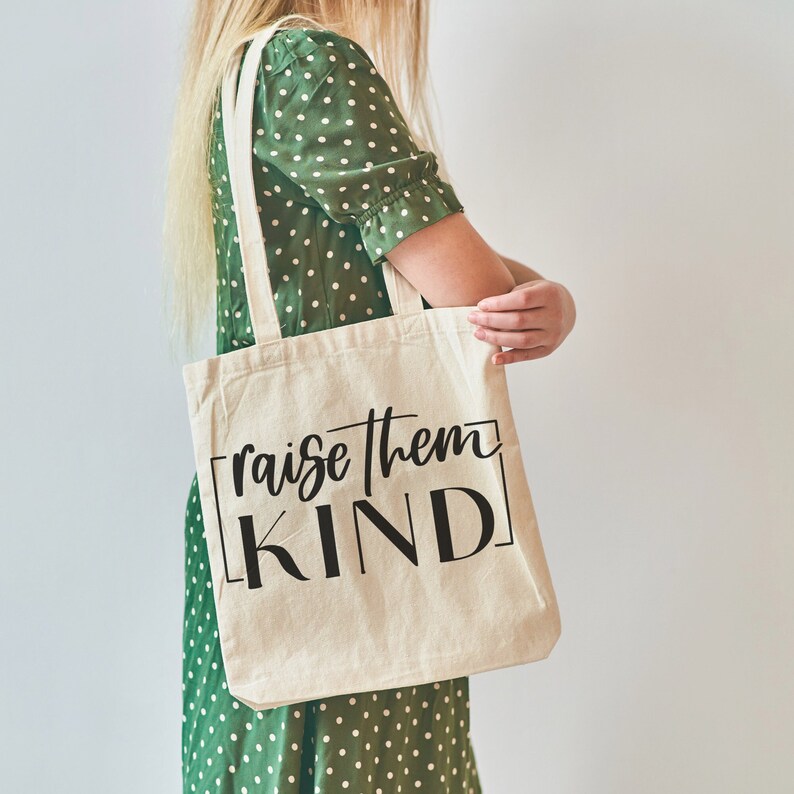 May include: A white tote bag with black text that reads "raise them KIND" in a script font. The bag is being held by a person wearing a green polka dot dress.