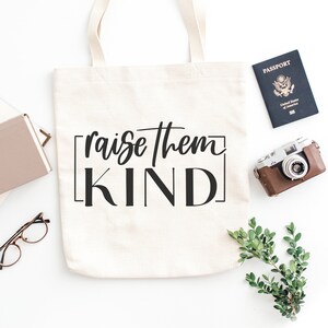 May include: A white canvas tote bag with black text that reads "raise them KIND" in a square frame. The bag is lying flat on a white surface with a passport, camera, and greenery in the background.