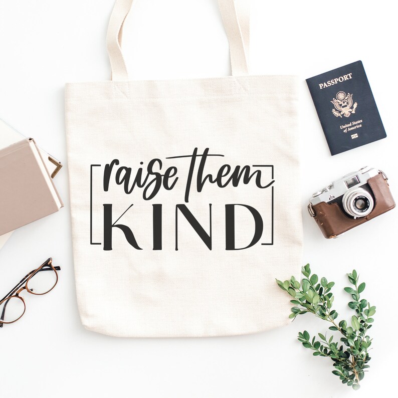 May include: A white canvas tote bag with black text that reads "raise them KIND" in a square frame. The bag is lying flat on a white surface with a passport, camera, and greenery in the background.