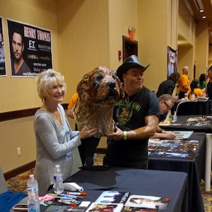 Puede incluir: Una gran escultura detallada de la cabeza de un perro con pelaje marrón y beige es sostenida por dos personas. La escultura tiene una apariencia realista. El fondo incluye mesas, carteles y otras personas.