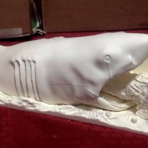 May include: A white sculpture depicting a shark attacking a person. The shark is open-mouthed, with the person holding a knife. The sculpture is on a white base, with a small box nearby.