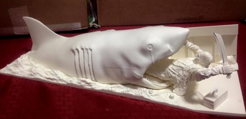 May include: A white sculpture depicting a shark attacking a person. The shark is open-mouthed, with the person holding a knife. The sculpture is on a white base, with a small box nearby.