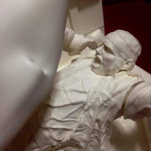 May include: A white sculpture of a person in a distressed pose, with a bandage on their head and an open mouth. The sculpture has detailed textures and is set against a neutral background.