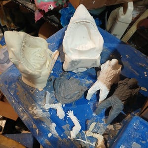May include: A collection of unfinished sculptures and molds, including a shark jaw, human figures, and other abstract shapes. The pieces are in various stages of completion, with a mix of white and gray materials. The items are arranged on a blue surface.