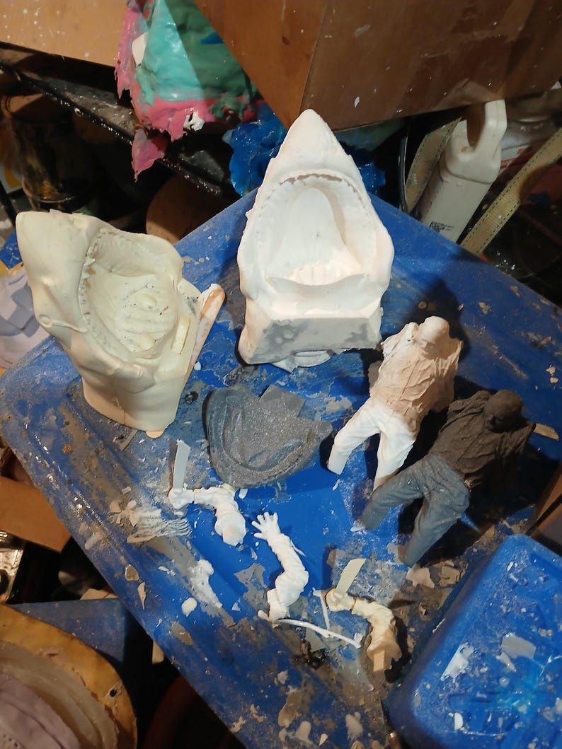May include: A collection of unfinished sculptures and molds, including a shark jaw, human figures, and other abstract shapes. The pieces are in various stages of completion, with a mix of white and gray materials. The items are arranged on a blue surface.