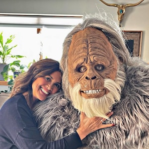 Könnte beinhalten: Eine Frau umarmt lächelnd ein großes Bigfoot-Kostüm. Das Kostüm hat ein detailliertes, realistisches Gesicht mit hellem Braunton, weißem Bart und grauem Fell. Die Frau trägt ein dunkelblaues Langarmshirt.