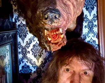 Könnte beinhalten: Ein Mann mit langen braunen Haaren steht vor einer Wand mit einer schwarz-weißen gemusterten Tapete. Ein ausgestopfter Tierkopf mit einem braunen und rosafarbenen Gesicht ist an der Wand hinter ihm angebracht.