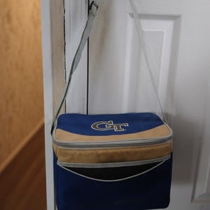 May include: A blue and tan insulated cooler bag with a shoulder strap. The bag features the letters "GT" in gold on the front. The cooler is hanging from a gold door knob.