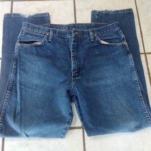 K&ouml;nnte beinhalten: Eine blaue Jeanshose aus Denim, flach hingelegt. Die Jeans hat ein klassisches F&uuml;nf-Taschen-Design mit Knopf- und Rei&szlig;verschluss. Der Denim hat ein verwaschenes Aussehen, mit sichtbaren Gebrauchsspuren. Die Jeans hat eine mittlere Waschung.