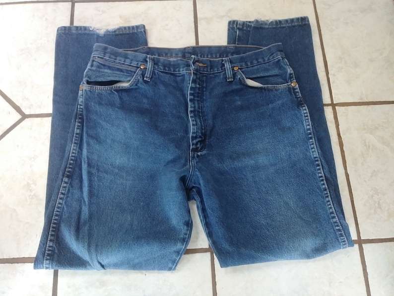 K&ouml;nnte beinhalten: Eine blaue Jeanshose aus Denim, flach hingelegt. Die Jeans hat ein klassisches F&uuml;nf-Taschen-Design mit Knopf- und Rei&szlig;verschluss. Der Denim hat ein verwaschenes Aussehen, mit sichtbaren Gebrauchsspuren. Die Jeans hat eine mittlere Waschung.