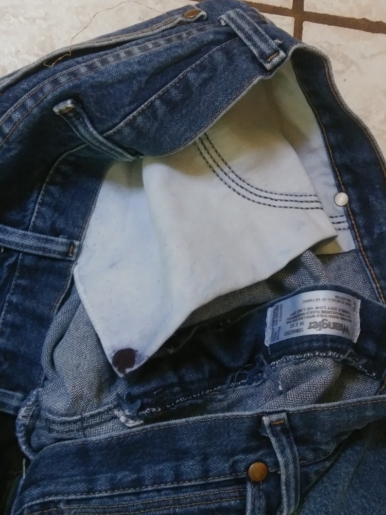 K&ouml;nnte beinhalten: Nahaufnahme einer blauen Jeans mit sichtbarem wei&szlig;em Taschenfutter. Die Jeans hat ein klassisches F&uuml;nf-Taschen-Design mit Knopf- und Rei&szlig;verschluss. Das Wrangler-Markenetikett ist sichtbar.
