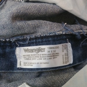 K&ouml;nnte beinhalten: Nahaufnahme einer blauen Wrangler-Jeans. Das Etikett lautet "Wrangler 13MWZDD 36 x 32" und enth&auml;lt Waschanweisungen. Die Jeans besteht aus 100 % Baumwolle.