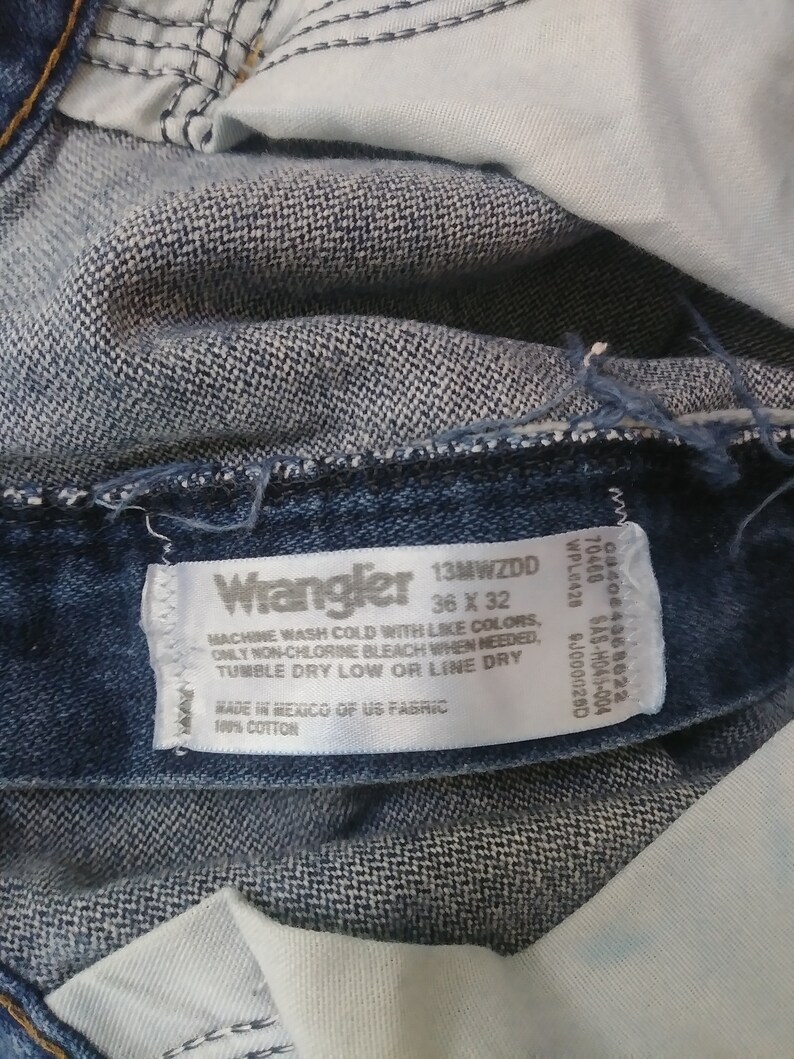 K&ouml;nnte beinhalten: Nahaufnahme einer blauen Wrangler-Jeans. Das Etikett lautet "Wrangler 13MWZDD 36 x 32" und enth&auml;lt Waschanweisungen. Die Jeans besteht aus 100 % Baumwolle.