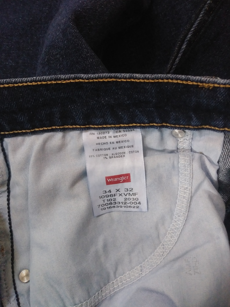 May include: A close-up of a Wrangler jeans label showing the size 34 x 32, style number 1096FXVMF, and other product details. The label is white with black text and a red Wrangler logo.