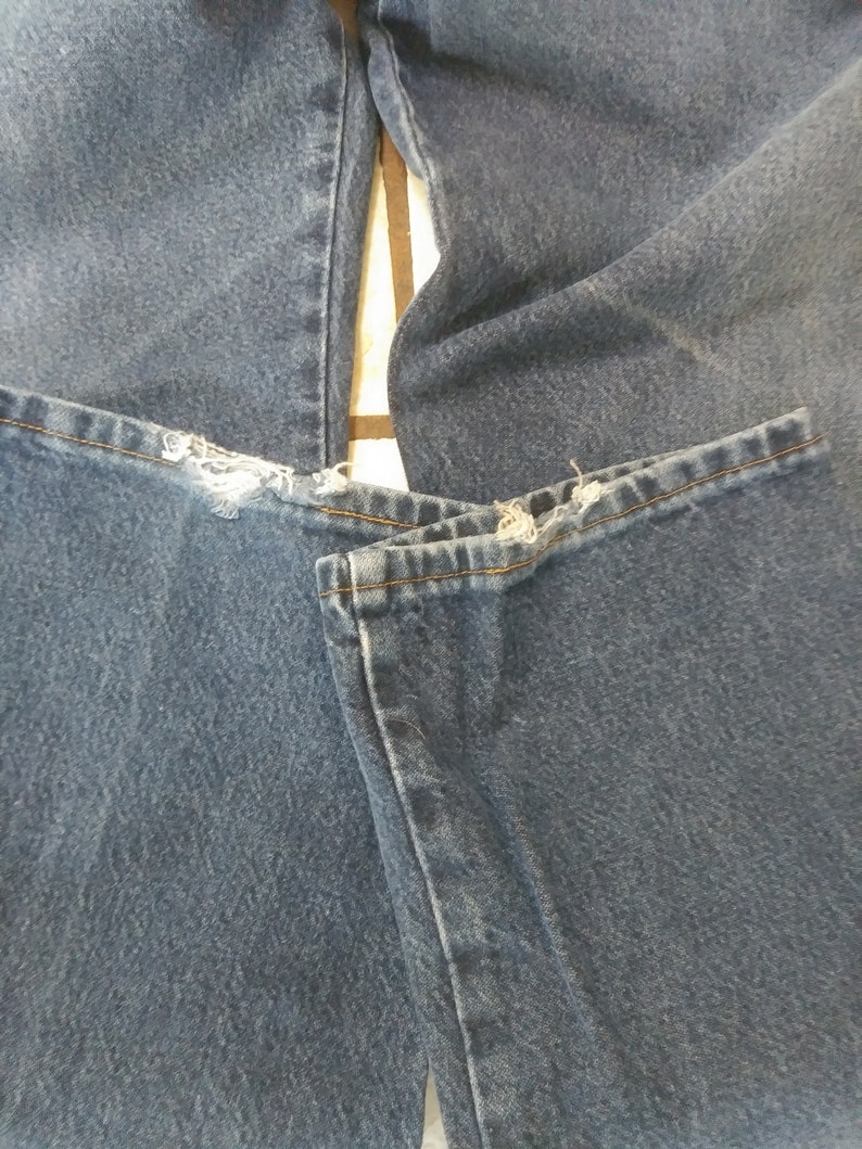 K&ouml;nnte beinhalten: Nahaufnahme einer blauen Jeans mit ausgefransten Details am Saum. Die Jeans ist gefaltet und zeigt die Textur und Farbe des Stoffes. Die N&auml;hte sind entlang der N&auml;hte sichtbar.
