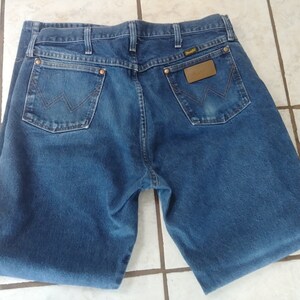 K&ouml;nnte beinhalten: Blaue Jeans aus Denim mit dem Wrangler-Logo auf der Ges&auml;&szlig;tasche und einem braunen Lederaufn&auml;her. Die Jeans hat zwei Ges&auml;&szlig;taschen und ein klassisches F&uuml;nf-Taschen-Design. Der Denim hat eine mittlere Waschung und die Jeans scheinen gefaltet zu sein.