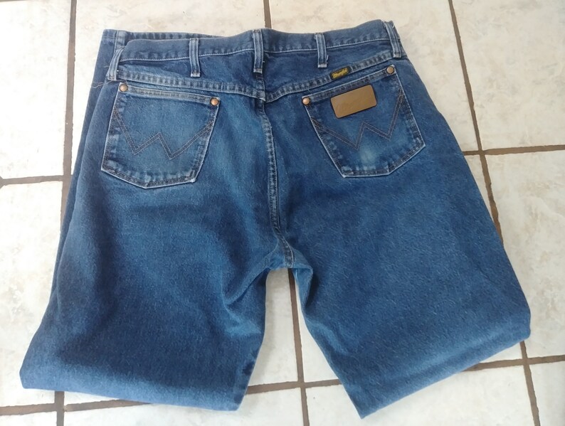 K&ouml;nnte beinhalten: Blaue Jeans aus Denim mit dem Wrangler-Logo auf der Ges&auml;&szlig;tasche und einem braunen Lederaufn&auml;her. Die Jeans hat zwei Ges&auml;&szlig;taschen und ein klassisches F&uuml;nf-Taschen-Design. Der Denim hat eine mittlere Waschung und die Jeans scheinen gefaltet zu sein.