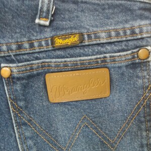 K&ouml;nnte beinhalten: Nahaufnahme der Ges&auml;&szlig;tasche einer blauen Jeans. Das Markenetikett Wrangler ist auf der Tasche und oberhalb der Tasche sichtbar. Die N&auml;hte sind in einer kontrastierenden Beiget&ouml;nung gehalten. Die Jeans hat ein klassisches F&uuml;nf-Taschen-Design.