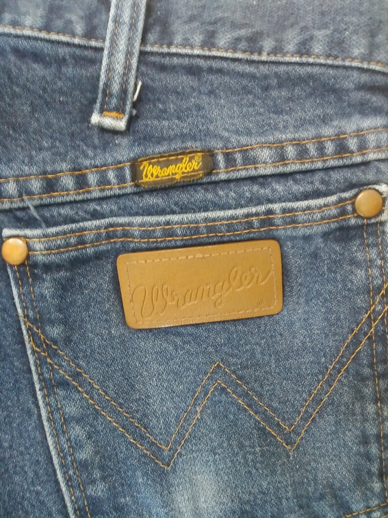 K&ouml;nnte beinhalten: Nahaufnahme der Ges&auml;&szlig;tasche einer blauen Jeans. Das Markenetikett Wrangler ist auf der Tasche und oberhalb der Tasche sichtbar. Die N&auml;hte sind in einer kontrastierenden Beiget&ouml;nung gehalten. Die Jeans hat ein klassisches F&uuml;nf-Taschen-Design.