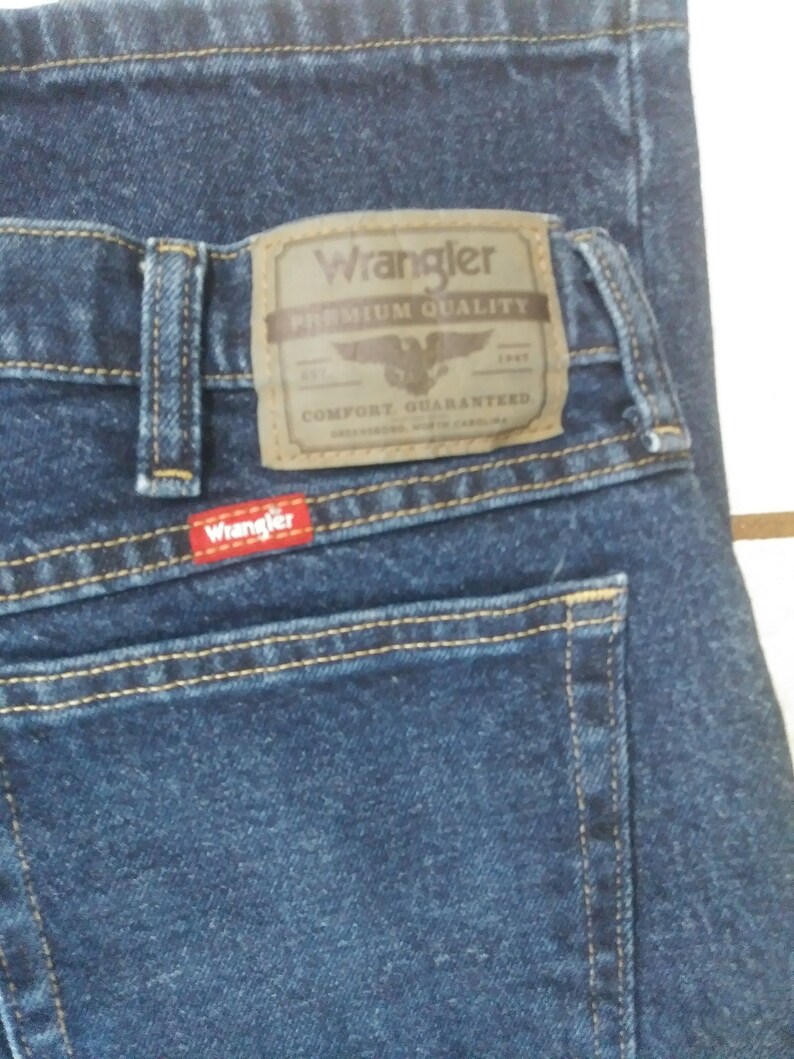 May include: A pair of blue denim jeans with a brown leather Wrangler label that says "Premium Quality" and "Comfort Guaranteed".