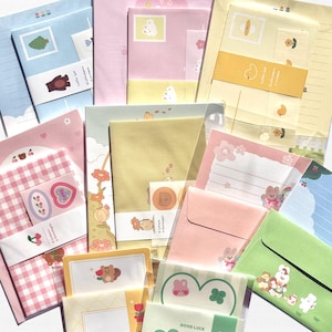 May include: A collection of stationery sets featuring various pastel-coloured envelopes and letter papers with printed illustrations of animals, flowers, and fruits. Each set is packaged in clear plastic, displaying patterns like gingham, solid colours, and decorative borders with small graphic icons.