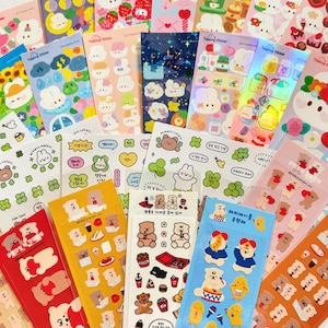 May include: A collection of multiple sticker sheets featuring various cartoon characters, including bears, rabbits, and chicks, alongside food items, lucky clovers, and decorative icons with Korean text and holographic finishes.
