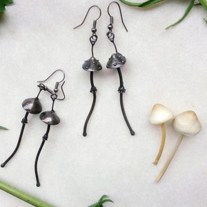 May include: Handmade silver mushroom earrings with a textured cap design and long, curved stems. The earrings are displayed with two real mushrooms and greenery, creating a whimsical, nature-inspired aesthetic. The earrings are a unique accessory.