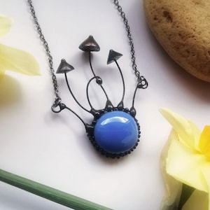 May include: A silver chain necklace with a blue glass pendant. The pendant is surrounded by black metal wire that is shaped like mushrooms.