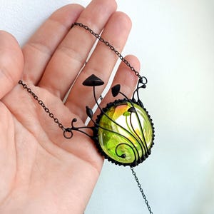 May include: A black metal necklace with a green glass pendant featuring a whimsical design of mushrooms and vines.