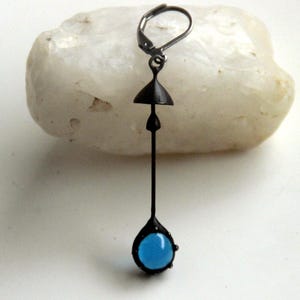 Puede incluir: Un pendiente negro con una piedra azul en forma de lágrima y un pequeño cono negro en la parte superior.