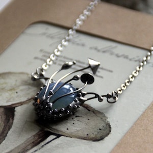 Può includere: Collana color argento con una pietra centrale ovale blu pallido. La pietra è incastonata in una montatura dettagliata e strutturata con piccoli accenti rotondi. Delicati elementi floreali stilizzati si estendono dal design centrale, collegati a una sottile catena.
