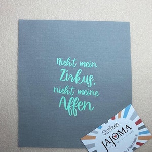 Könnte beinhalten: Ein grauer Stoffquadrat mit dem Text "nicht mein Zirkus, nicht meine Affen" in türkisfarbener Schrift. Eine Visitenkarte für Stofferia JAJOMA befindet sich in der rechten unteren Ecke.