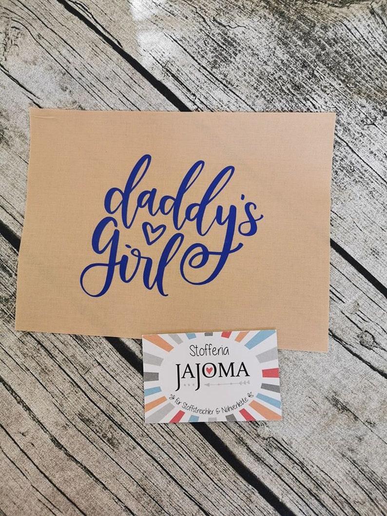 Puede incluir: Un cuadrado de tela marr&oacute;n con el texto "daddy's girl" en letras azules con dos peque&ntilde;os corazones rojos. Una peque&ntilde;a etiqueta colorida con el texto "Stoffena JAJOMA" est&aacute; en la esquina inferior derecha.
