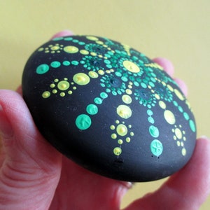 May include: A smooth, round, black stone featuring a hand-painted mandala design. The pattern consists of numerous small, raised dots in varying shades of bright lime green, emerald green, and pale yellow, arranged in a radial, symmetrical formation.
