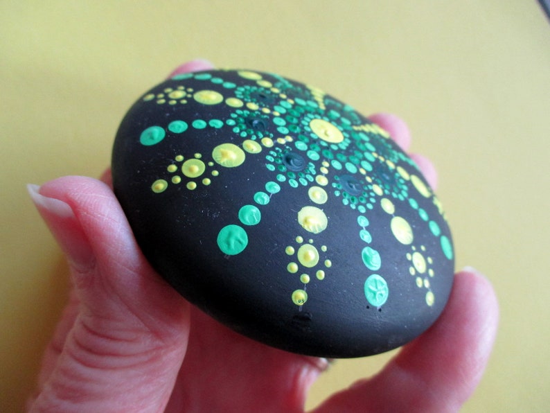 May include: A smooth, round, black stone featuring a hand-painted mandala design. The pattern consists of numerous small, raised dots in varying shades of bright lime green, emerald green, and pale yellow, arranged in a radial, symmetrical formation.