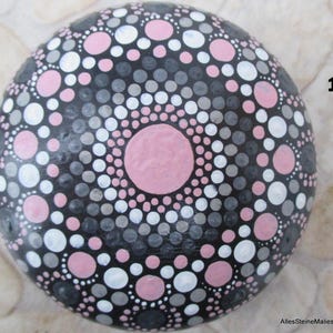 May include: A round, smooth stone painted with a mandala pattern of concentric circles in shades of pink, white, gray, and black. The design features a large central pink dot surrounded by alternating rings of smaller dots. The number 1 is visible.