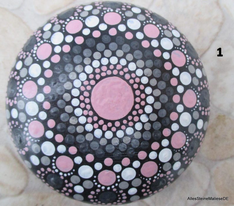May include: A round, smooth stone painted with a mandala pattern of concentric circles in shades of pink, white, gray, and black. The design features a large central pink dot surrounded by alternating rings of smaller dots. The number 1 is visible.