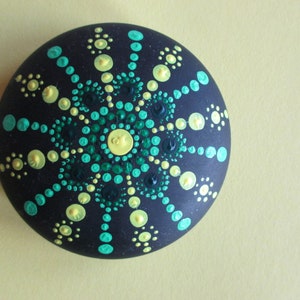 May include: A round, dark charcoal-colored stone featuring a hand-painted mandala design. The pattern consists of concentric circles of raised paint dots in shades of lime green, turquoise, and dark forest green, radiating from a central yellow dot.