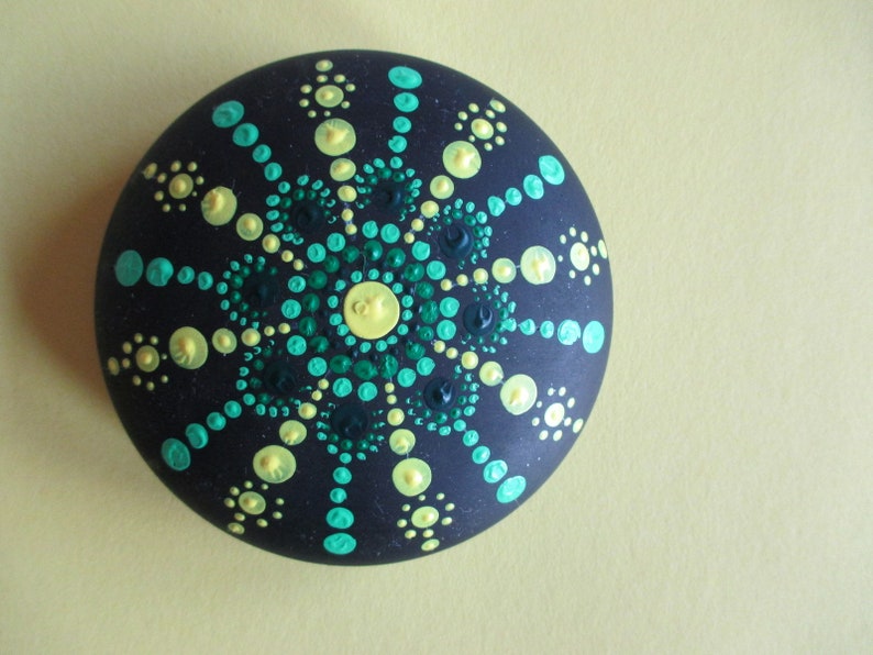 May include: A round, dark charcoal-colored stone featuring a hand-painted mandala design. The pattern consists of concentric circles of raised paint dots in shades of lime green, turquoise, and dark forest green, radiating from a central yellow dot.