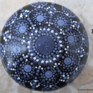 May include: A round, black stone featuring a hand-painted mandala design composed of numerous dots in varying shades of blue and white. The pattern radiates from a central blue dot, creating concentric circles. The number 3 and AllesSteineMalieseDE are visible.