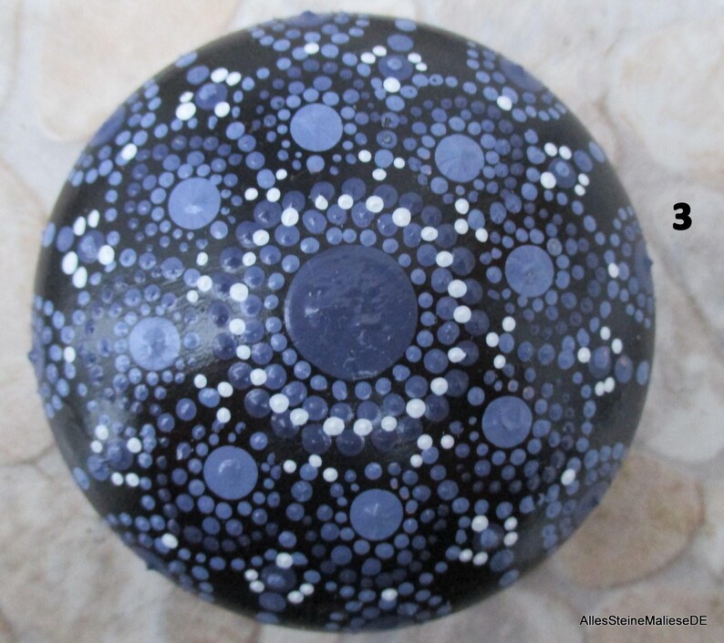 May include: A round, black stone featuring a hand-painted mandala design composed of numerous dots in varying shades of blue and white. The pattern radiates from a central blue dot, creating concentric circles. The number 3 and AllesSteineMalieseDE are visible.
