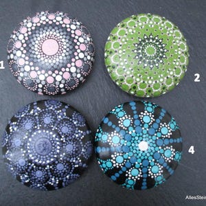 May include: Four round stones decorated with intricate dot mandala patterns. Stone 1 features pink, gray, and white dots. Stone 2 has green and white dots. Stone 3 displays purple and white dots. Stone 4 shows blue, teal, and white dots. Text reads AllesSteineMalieseDE.