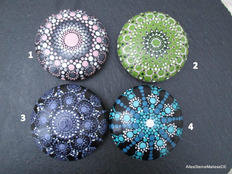 May include: Four round stones decorated with intricate dot mandala patterns. Stone 1 features pink, gray, and white dots. Stone 2 has green and white dots. Stone 3 displays purple and white dots. Stone 4 shows blue, teal, and white dots. Text reads AllesSteineMalieseDE.