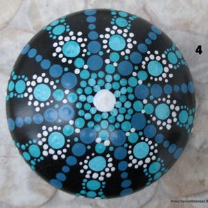 May include: A round black stone painted with a mandala pattern consisting of numerous dots in shades of turquoise, light blue, dark blue, and white. The design radiates from a single white center dot. The number 4 is visible on the right side.